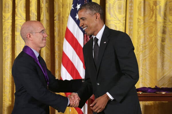 President Barack Obama shaking hands with Mr. Katzenberg, who has a medal on a purple ribbon around his neck. Both are wearing dark suits and standing in front of a gold curtain and an American flag. 